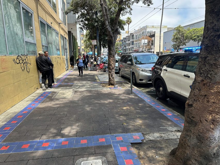 A city sidewalk with people walking, parked cars on the right, trees lining the street, and a tiled border along the pavement.