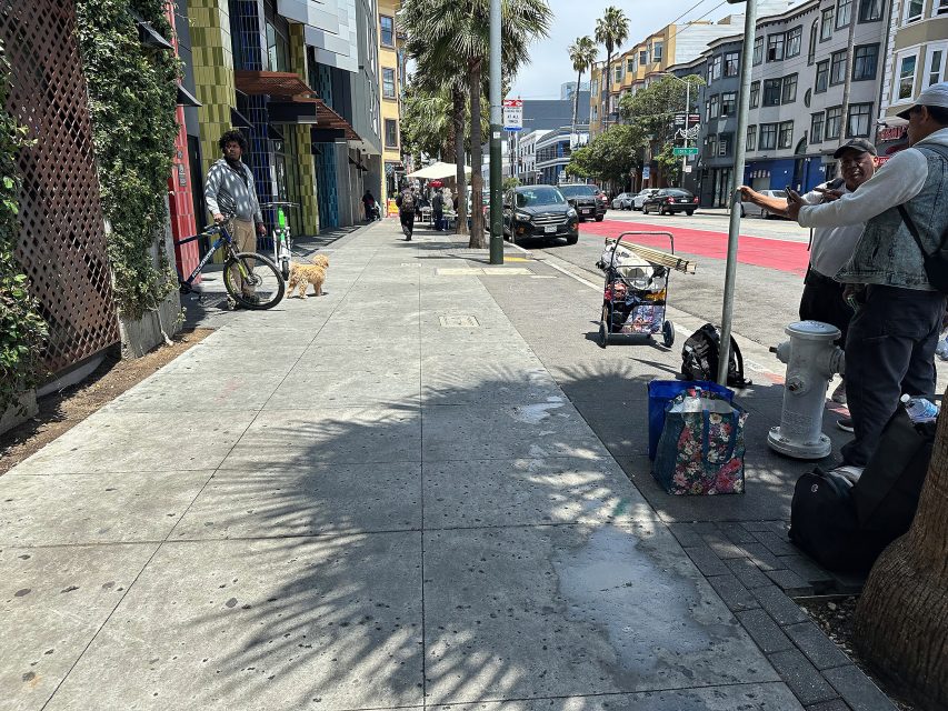 A city sidewalk with people standing and sitting, a man with a bicycle and dog, parked cars, and buildings lining the street on a sunny day.