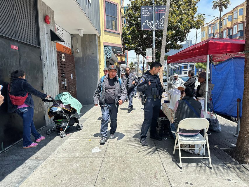 Two police officers stand near a table with people seated under a red canopy on a busy city sidewalk; pedestrians walk by and colorful buildings line the street.