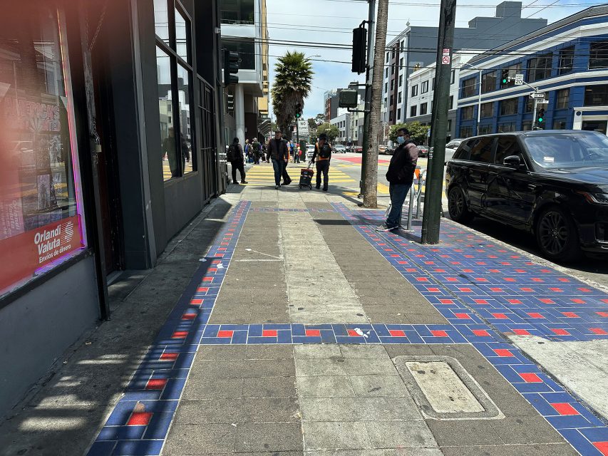 People walk along a city sidewalk decorated with blue and red tile patterns, with cars parked at the curb and buildings lining the street in the background.
