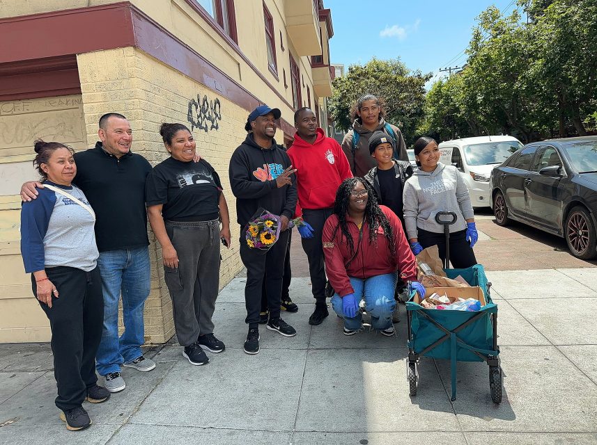A group of people pose for a photo on a city sidewalk with a cart containing supplies, wearing gloves and casual clothes. Some graffiti is visible on the building wall behind them.