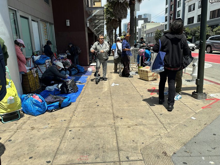 People are gathered on a city sidewalk with bags and belongings, some sitting or standing near a building, while others walk along the street on a sunny day.
