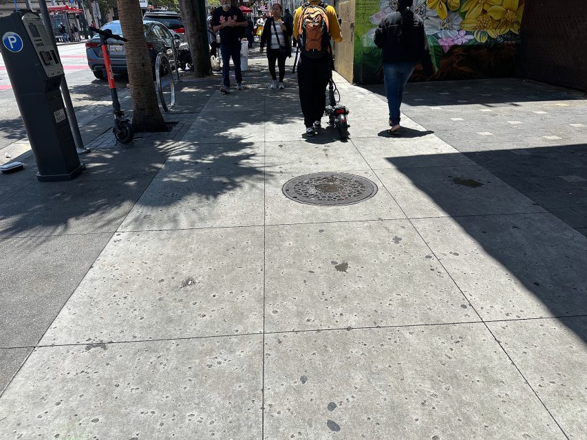 People walk on a sunny city sidewalk near parked scooters and a mural, with shadows from nearby trees visible on the pavement.