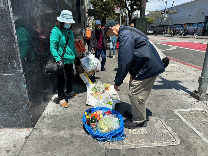Two people wearing masks examine goods laid out on the sidewalk as street vendors sell food and drinks on a city street corner.