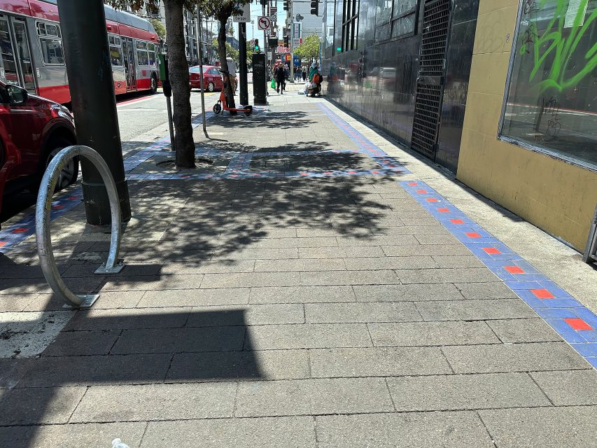 A city sidewalk in sunlight with blue and red tile borders, a bike rack, trees, a parked bus, pedestrians, and buildings alongside.