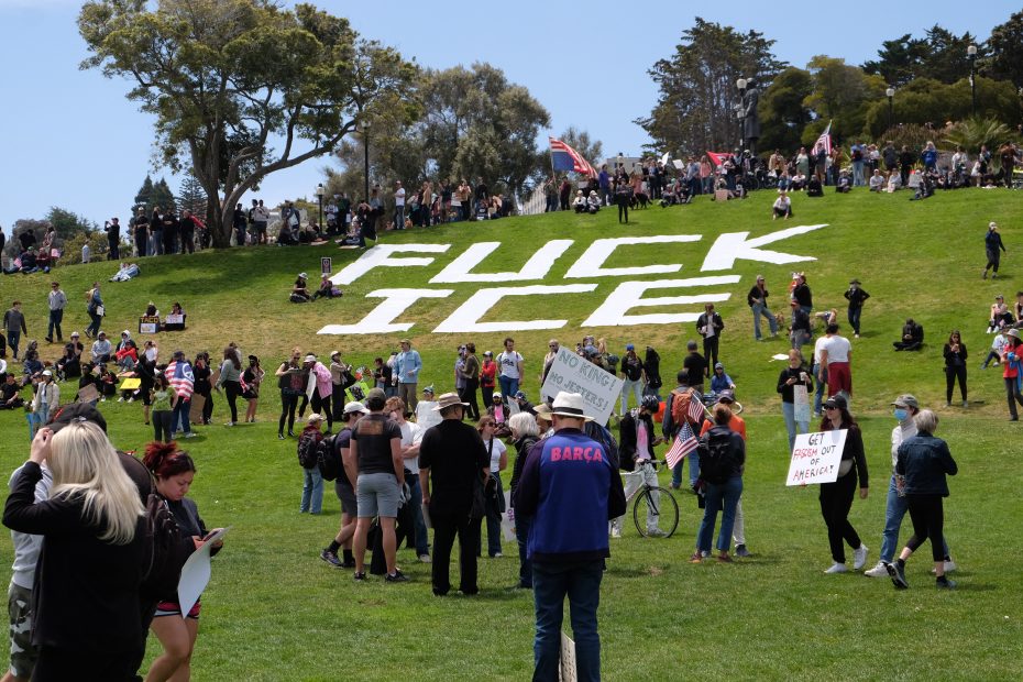 A crowd gathers in a park near a hillside with large white letters spelling "FUCK ICE," joining the protest as people hold signs and socialize on the grass.