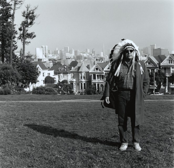 A man wearing a feathered headdress and coat stands in a park with Victorian houses and a city skyline in the background.