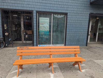 An orange wooden bench with "sfbabc.org" text sits on a sidewalk in front of a blue building with windows and doors.