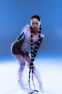 A person poses under blue lighting, wearing a lace-up dress, long braided hair extensions with bows, tall boots, and transparent gloves.