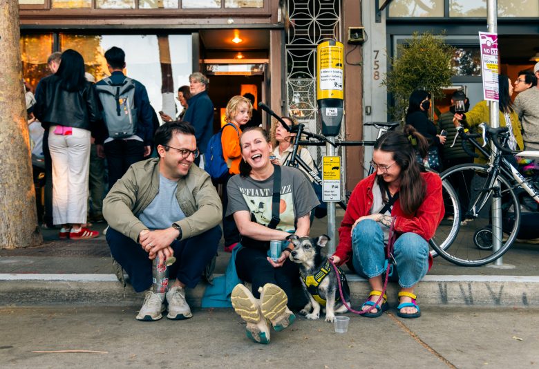 Three people sit on a sidewalk outside a busy storefront, smiling and talking with a small dog in front of them; bicycles and a crowd are visible in the background.