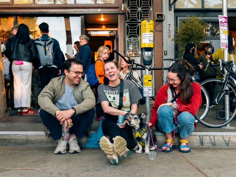 Three people sit on a sidewalk outside a busy storefront, smiling and talking with a small dog in front of them; bicycles and a crowd are visible in the background.