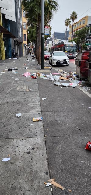 A city sidewalk littered with trash and debris, with a white car parked near a dumpster and palm trees lining the street.
