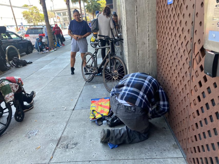 A person in a plaid shirt kneels on a city sidewalk near a wall, while others stand and walk nearby; bicycles are parked along the street.