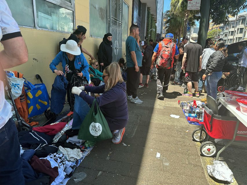 A group of people gather on a city sidewalk, some sorting through clothes on the ground, while others stand or sit nearby with bags and carts.