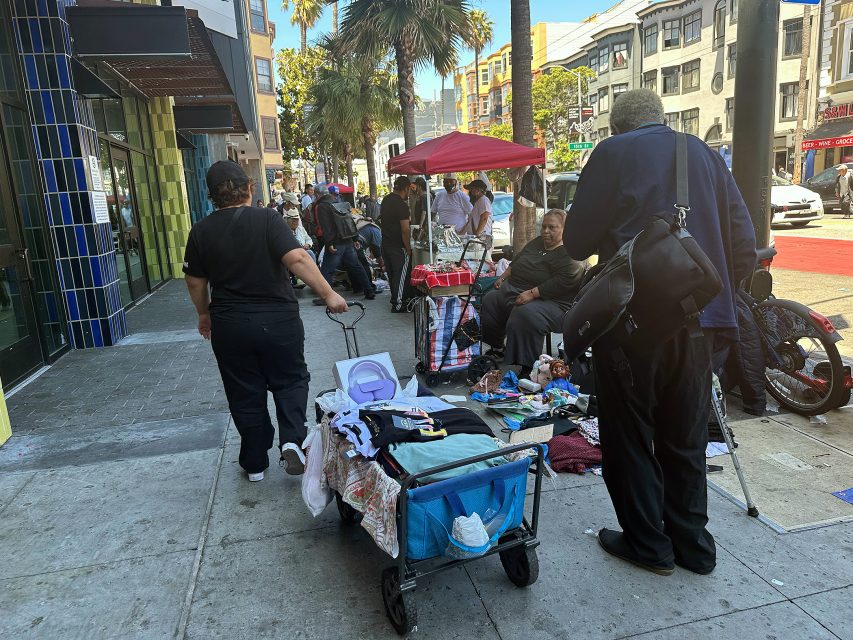 People gather on a city sidewalk with tables and carts displaying various items for sale or exchange; buildings and palm trees are visible in the background.