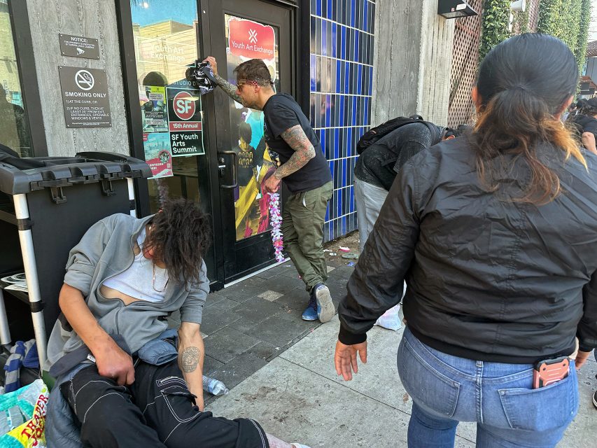 Several people are gathered outside a building entrance; one person sits slouched against a trash bin, while others stand or walk nearby on the sidewalk.