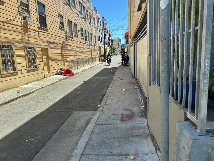 A narrow urban alley with people sitting along the right wall, one person lying on the left, and graffiti visible on the sidewalk and walls.