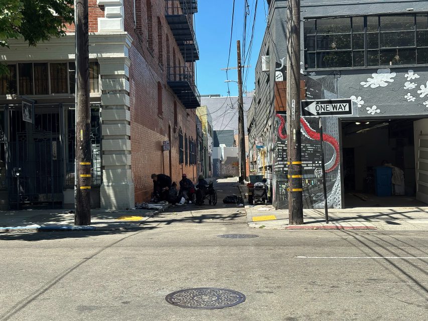 A group of people gather in the shade of an alley between brick and painted buildings; a one-way street sign is visible in the foreground.