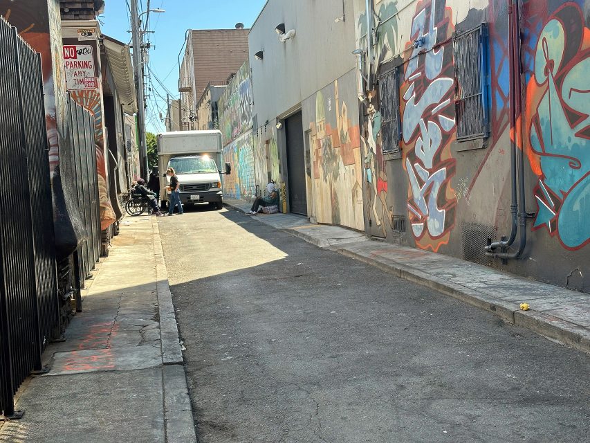 A narrow alley with graffiti-covered walls, a white truck parked, two people standing beside it, and another person sitting on the ground farther down.