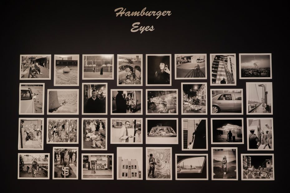 A wall display titled "Hamburger Eyes" features 24 black-and-white photographs arranged in a grid, depicting various urban scenes and portraits.