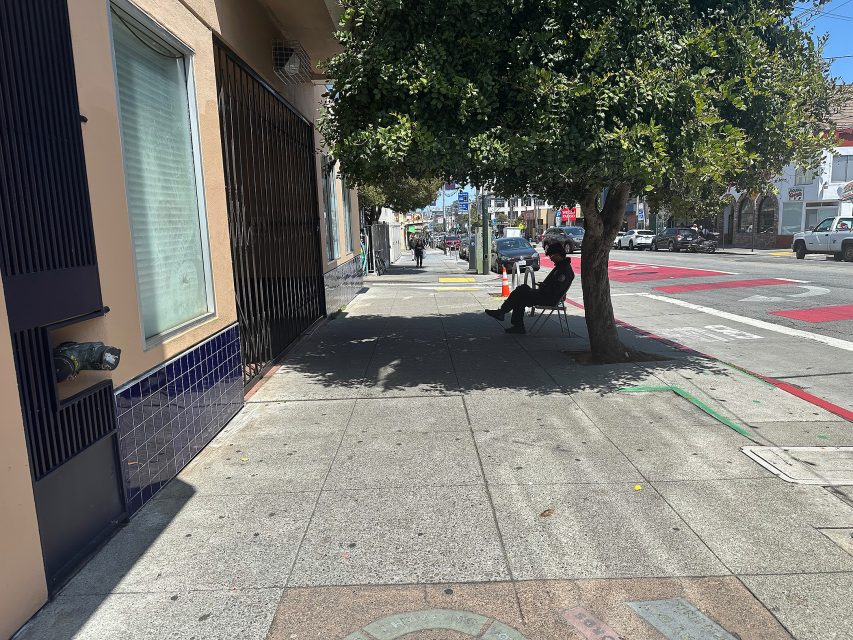 A person sits on a chair under a tree's shade on a city sidewalk near a street with cars and red bus lanes.