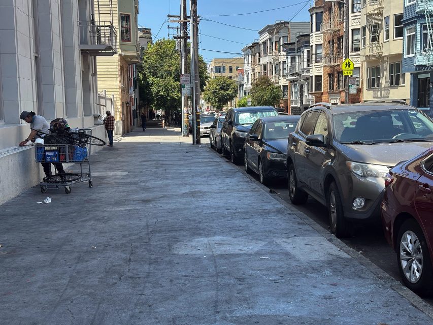 A person with a shopping cart stands by a building on a city sidewalk; parked cars line the street on a sunny day.