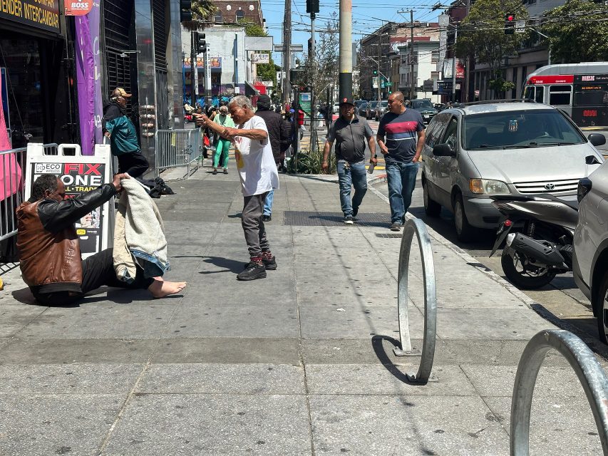 A man photographs another person sitting on the sidewalk near a storefront, while pedestrians walk by and cars are parked along the street.