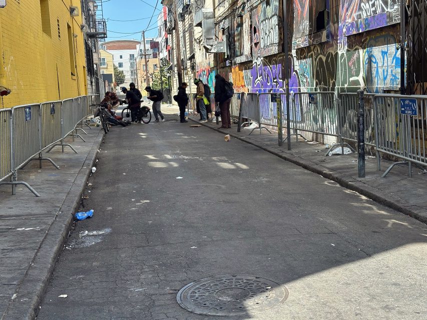 A narrow alley with graffiti-covered walls, metal barricades along the sides, and several people gathered or sitting in the distance. Trash is scattered on the ground.