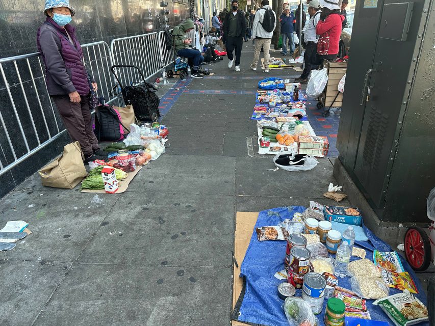 People set up food and groceries for sale on the sidewalk in an urban area; some are standing and others are seated nearby.