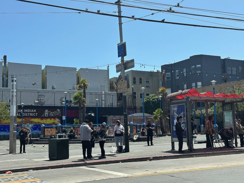 People wait at a bus stop and socialize in a city plaza with murals, palm trees, and buildings in the background under a clear sky.