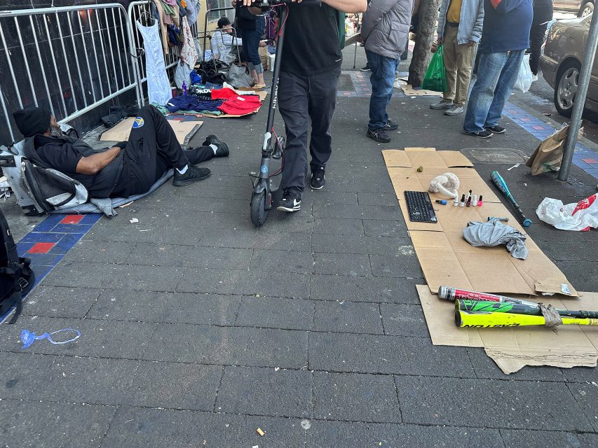 A person rides an electric scooter past a person lying on the sidewalk and items displayed on cardboard, including a keyboard, nail polish, and a cricket bat.