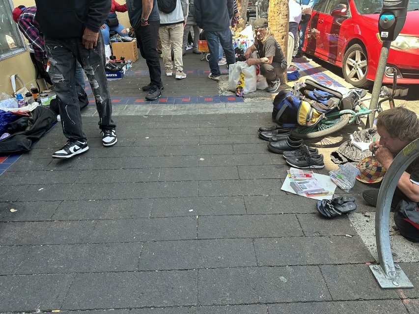 People are gathered on a city sidewalk near parked cars. Belongings are scattered, and a person sits on the ground reading and eating, surrounded by bags and personal items.