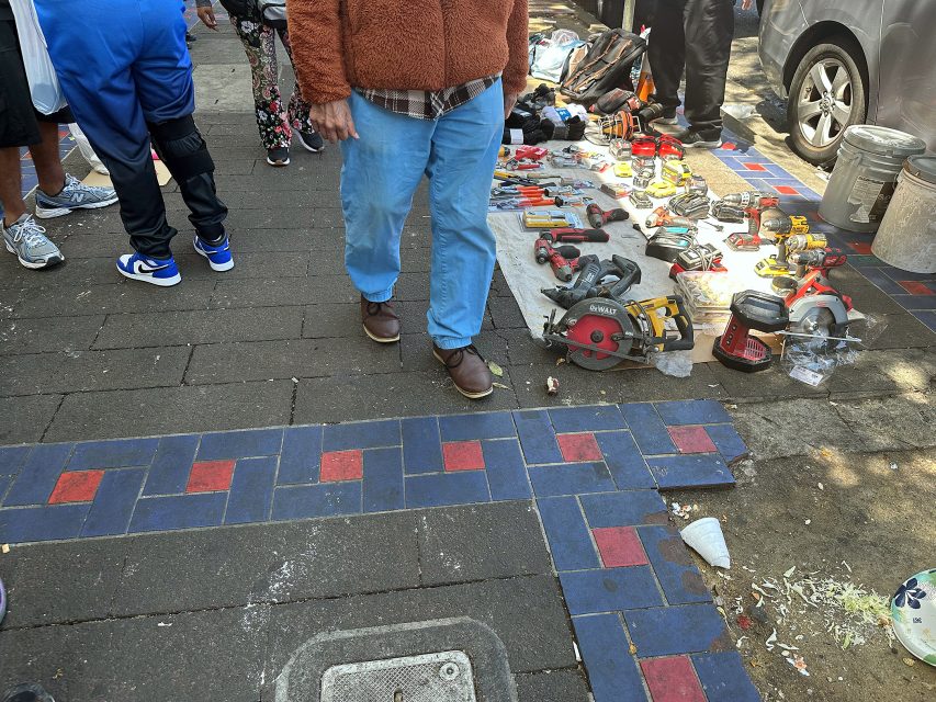 A person stands near a sidewalk display of various power tools and equipment laid out on a cloth at an outdoor market.
