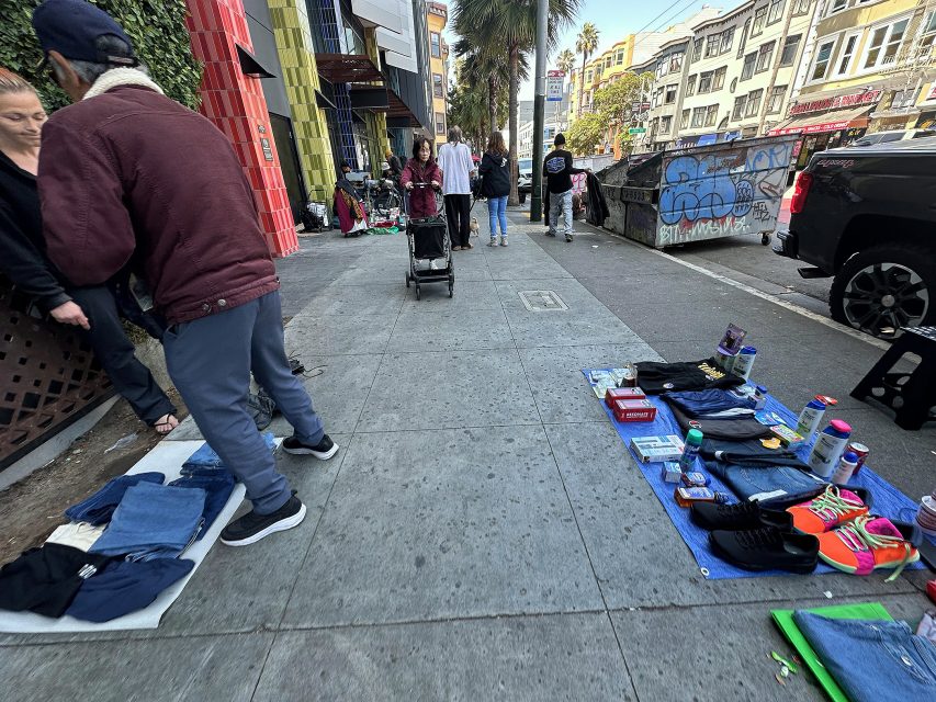 People selling clothes and various items on a city sidewalk; pedestrians and buildings are visible in the background.