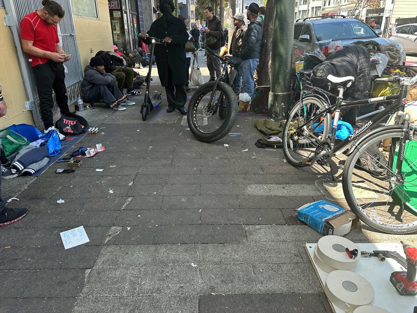 A group of people gather on a city sidewalk with bikes, belongings, and scattered items; some are sitting or standing near a tree and building.