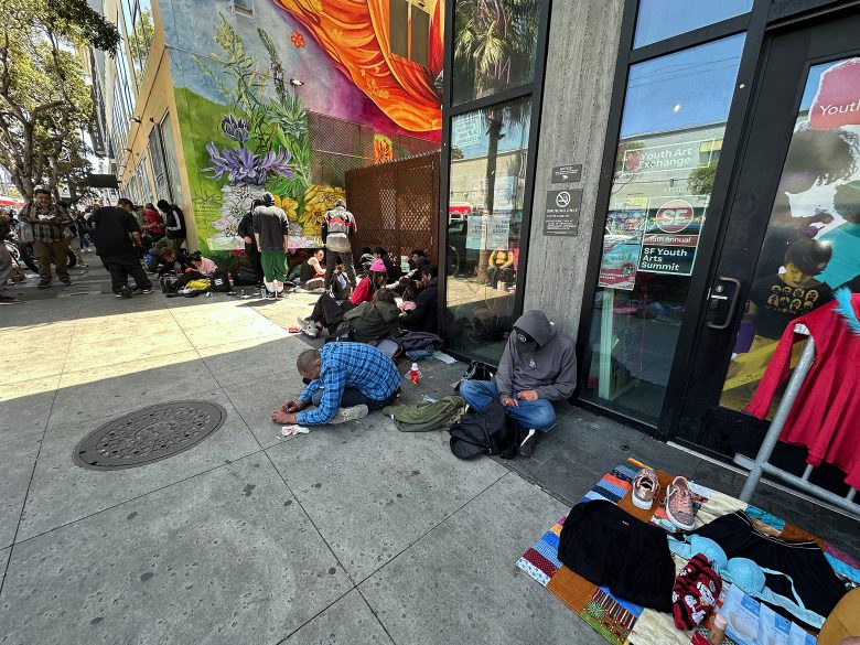 Several people sit or lie on a city sidewalk near a building with a colorful mural and storefront windows, with clothing and personal belongings nearby.