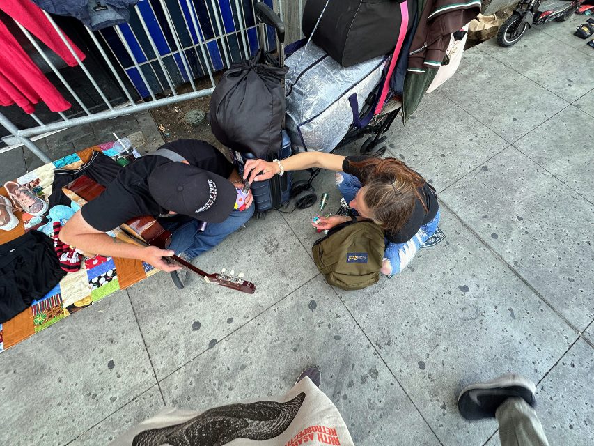 Two people sit on a city sidewalk near bags and belongings; one plays a guitar while the other reaches toward the guitar player with a small object in hand.