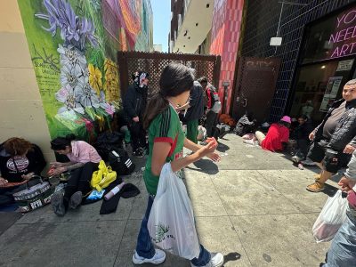 A person carrying plastic bags walks past people sitting on a city sidewalk near a colorful mural and storefront.