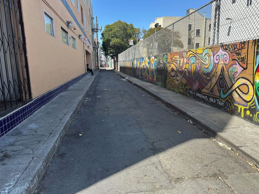 Narrow urban alley with graffiti-covered wall on the right, apartment building on the left, and a few people visible in the distance under a clear blue sky.