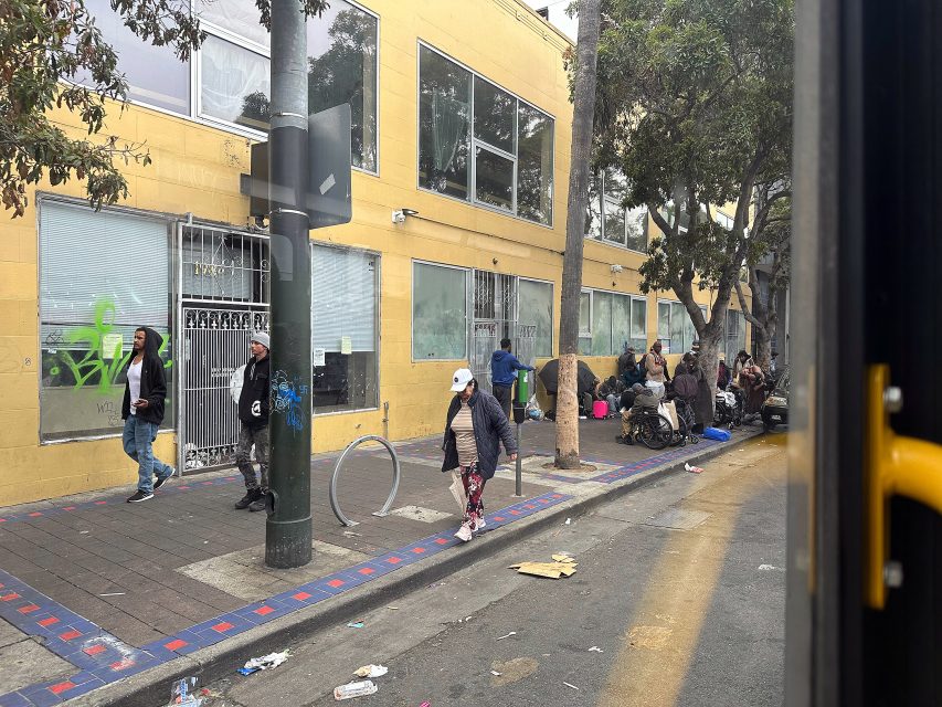 People walk and sit along a city sidewalk lined with trees and a yellow building, with scattered litter visible on the ground.