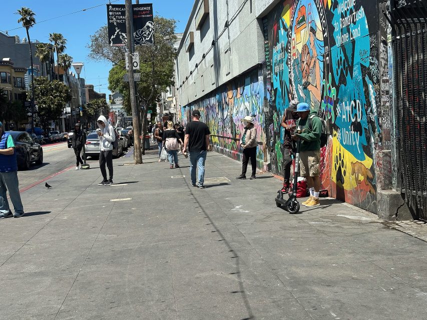 People walk along a city sidewalk next to a colorful graffiti-covered wall on a sunny day. One person stands near the wall with a scooter.