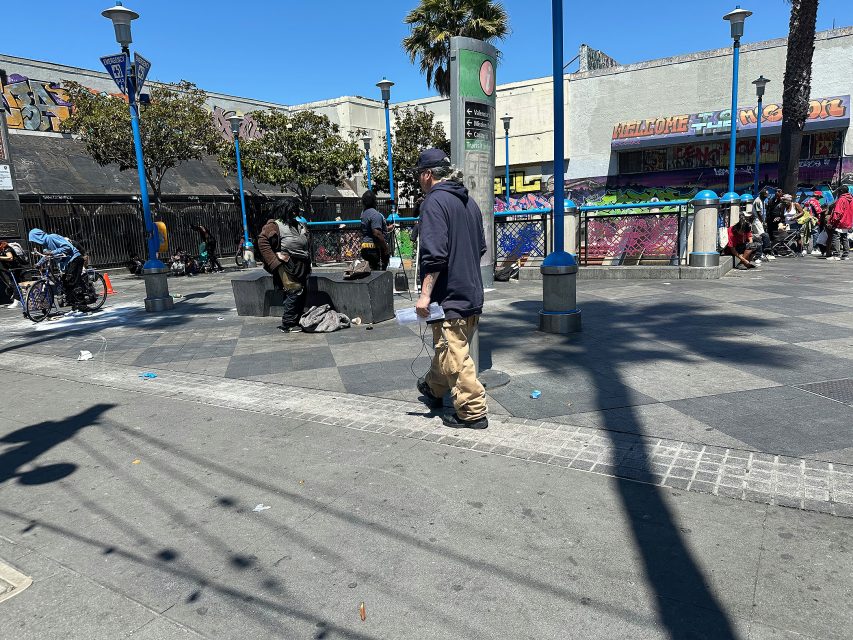 A man walks through a city plaza with graffiti-covered walls, while several people gather or sit nearby on a sunny day.