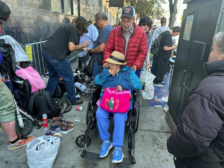 An elderly person in a wheelchair surrounded by people and belongings on a city sidewalk; a man in a red jacket stands behind her.