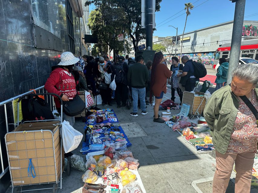 People gather on a city sidewalk where various goods, including snacks and household items, are displayed on tarps for sale.