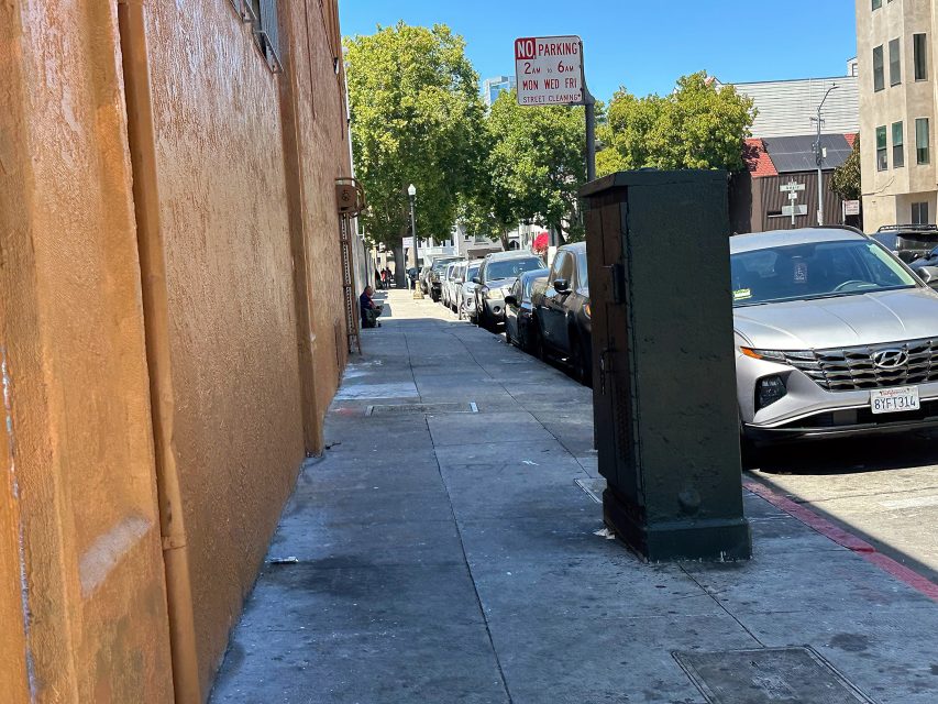 A city sidewalk with a row of parked cars, an orange building on the left, a utility box in the foreground, and a "No Parking" sign ahead under a clear blue sky.
