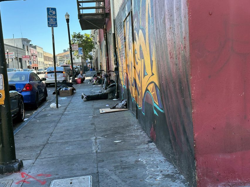 People sit and lie on a city sidewalk near a building with graffiti, while cars are parked along the street under a handicapped parking sign.
