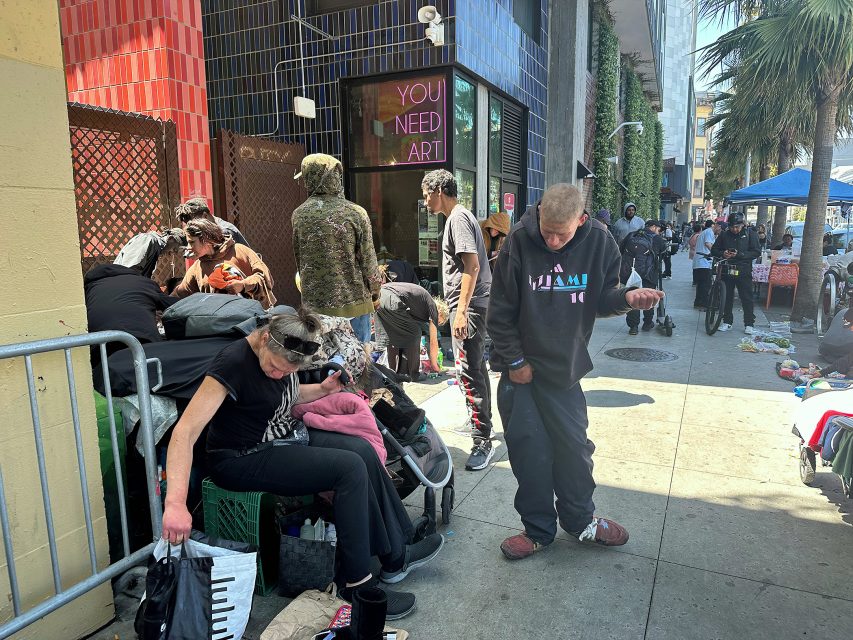 A group of people gather on a city sidewalk with belongings and tents; some sit or stand, while one man looks down at his hand. A neon "YOU NEED ART" sign is visible in the background.