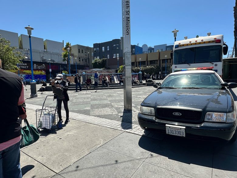 Police car and emergency vehicle parked on a city street as people walk and gather in an urban plaza during daytime.