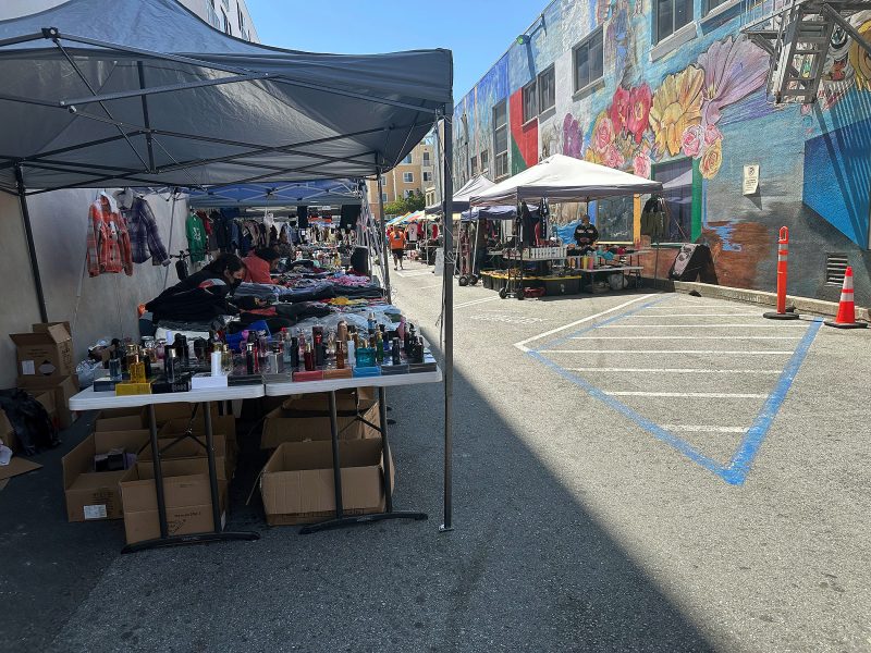 Outdoor market with tables displaying various goods, including perfumes and clothing, set up under tents along a mural-decorated alley on a sunny day.