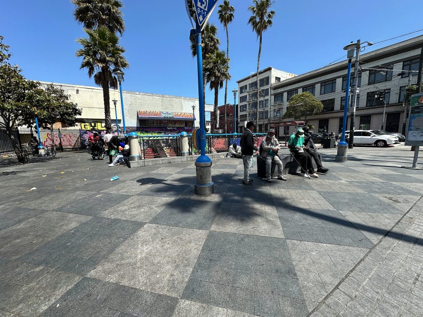 People are sitting on benches and standing in an urban plaza with palm trees, graffiti, and buildings visible in the background under a clear blue sky.